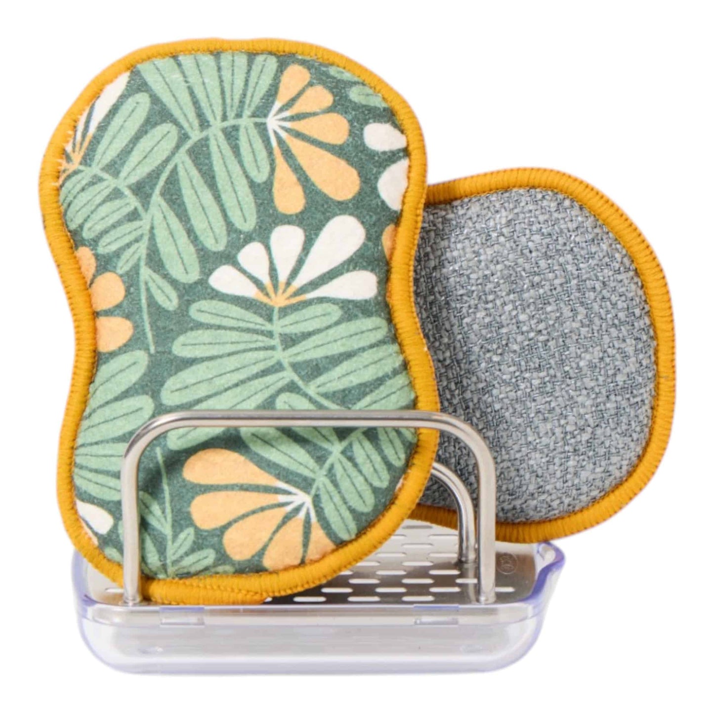 Two oval-shaped dishcloths with a green tropical leaf pattern and yellow-orange trim, shown with a gray textured backing and a clear plastic holder on a white background.