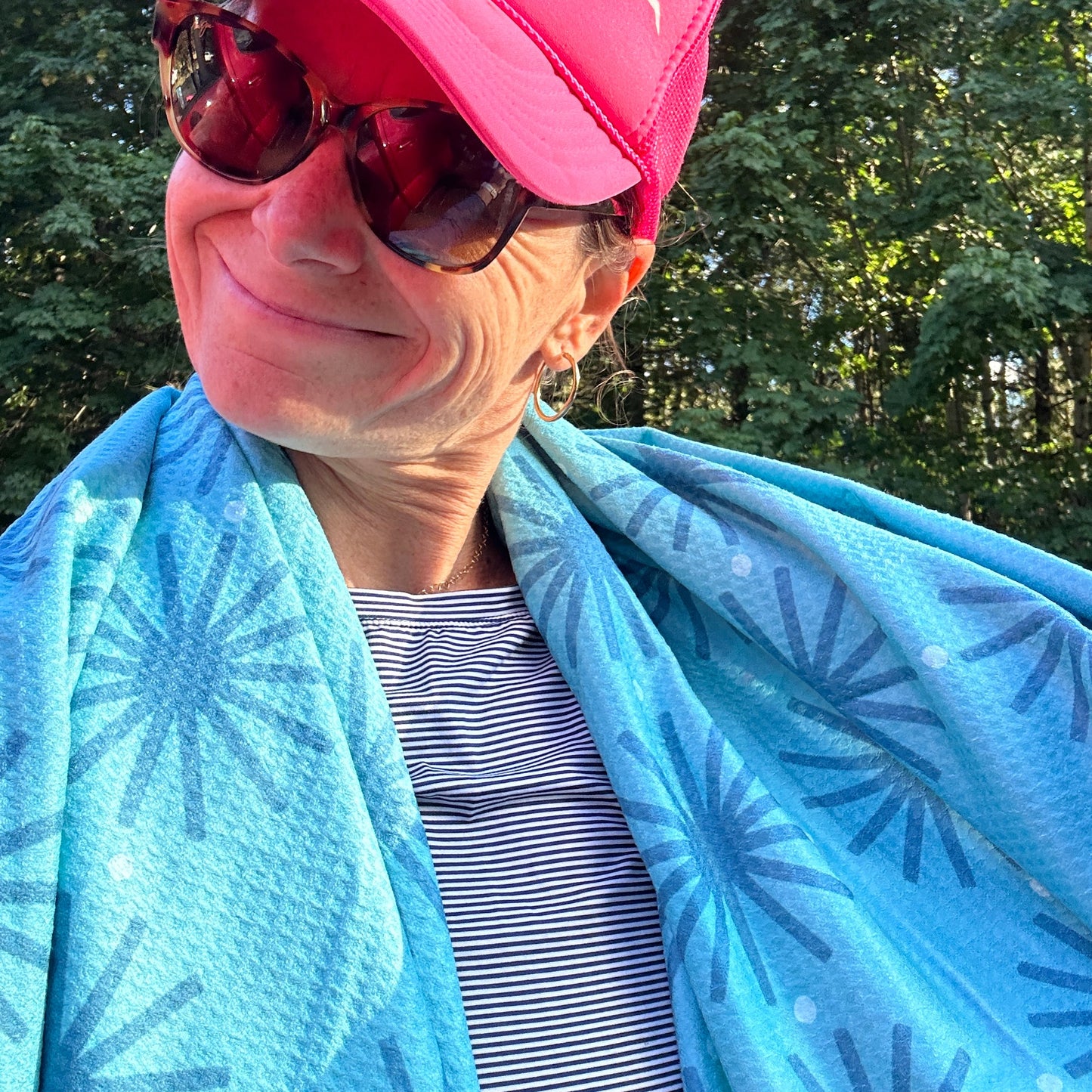 Person wearing a pink cap and sunglasses, wrapped in a blue patterned beach towel with trees in the background.