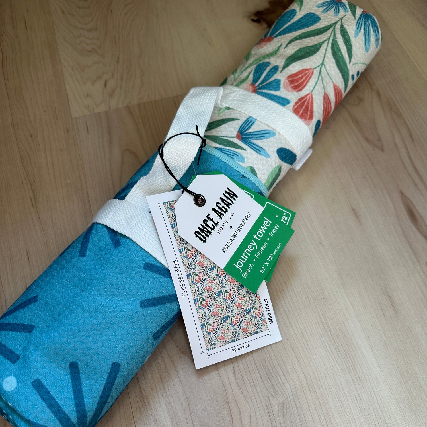 Rolled-up towel with floral and abstract patterns on a wooden floor, featuring a tag.