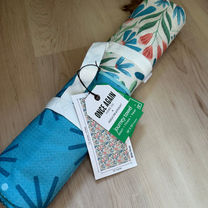 Rolled-up towel with floral and abstract patterns on a wooden floor, featuring a tag.