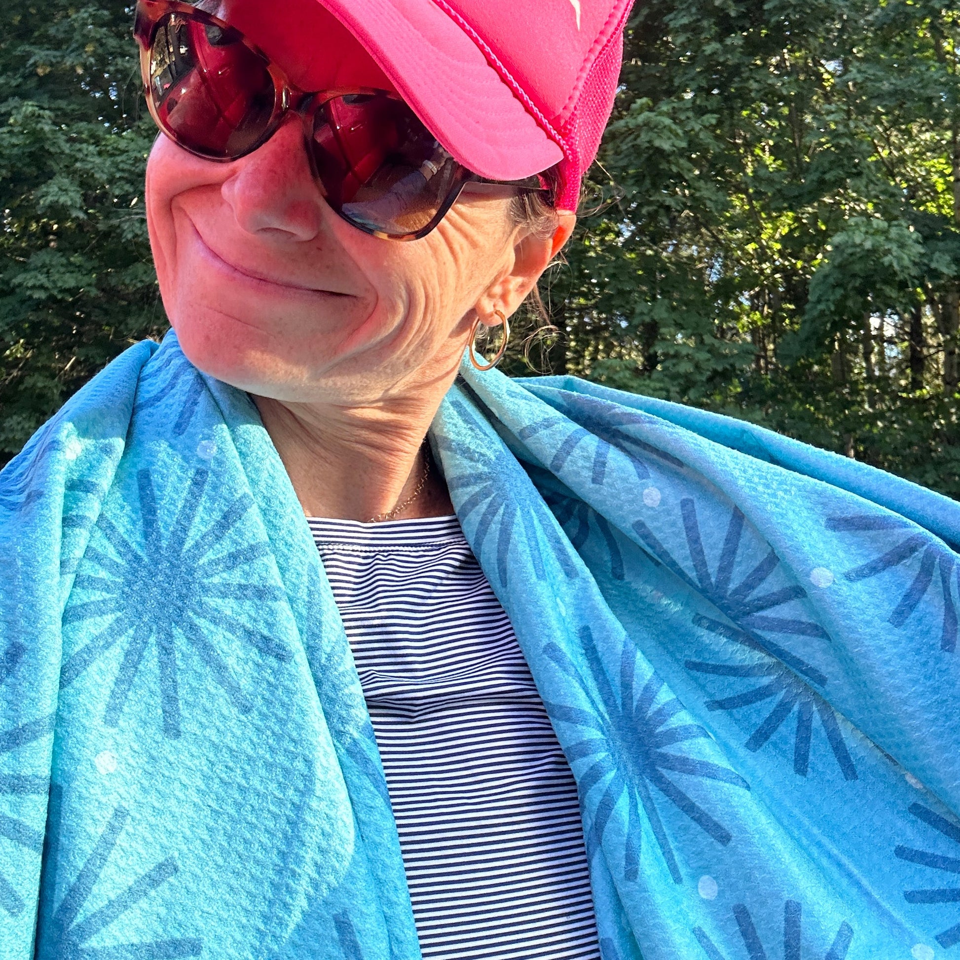 Person wearing a pink cap and sunglasses, wrapped in a blue patterned beach towel with trees in the background.