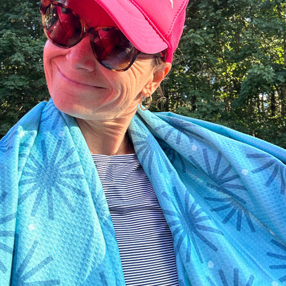 Person wearing a pink cap and sunglasses, wrapped in a blue patterned beach towel with trees in the background.